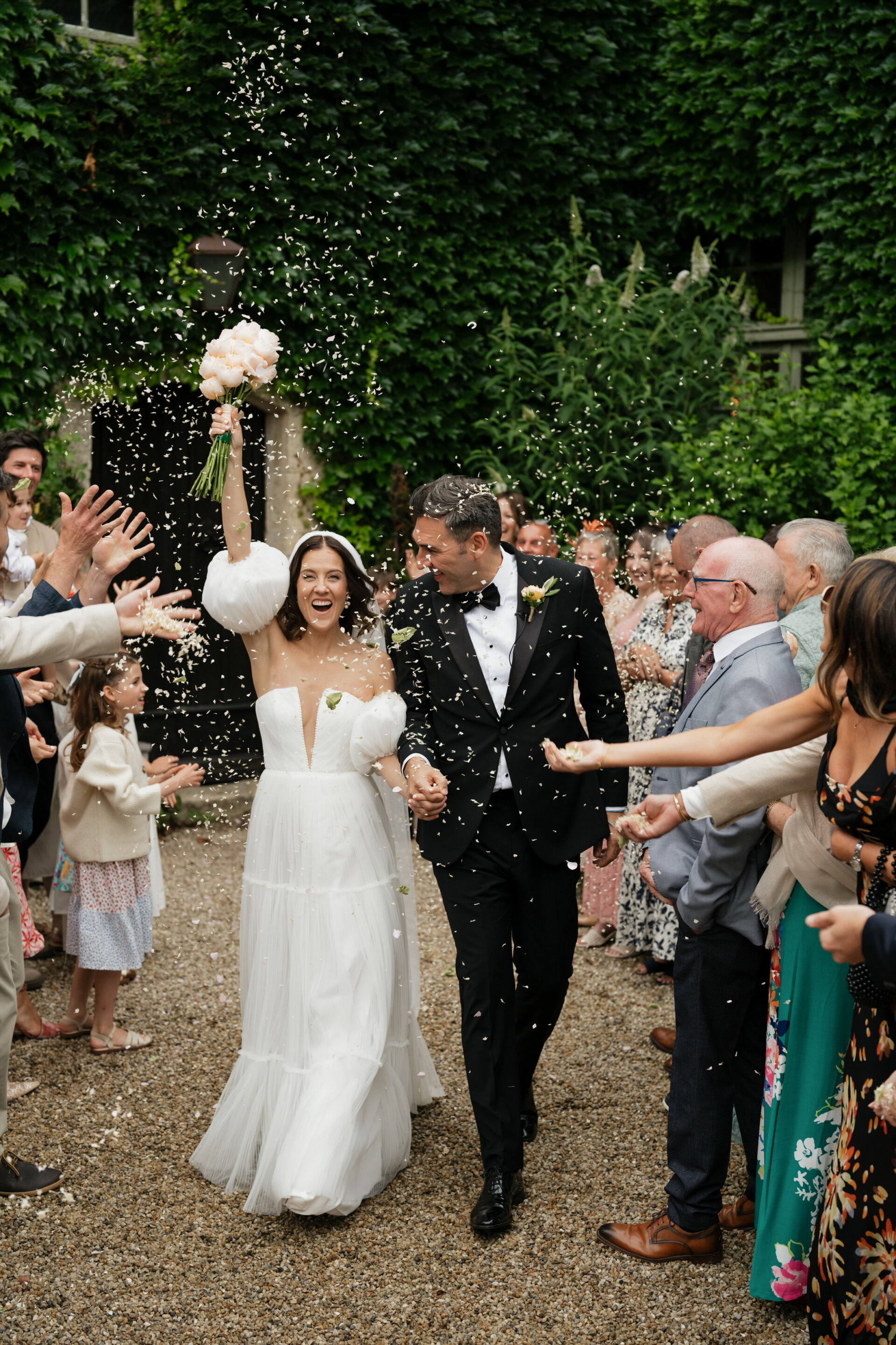 A bride and groom walk hand in hand through a crowd of guests who are throwing confetti, with lots of trees and plants around them.