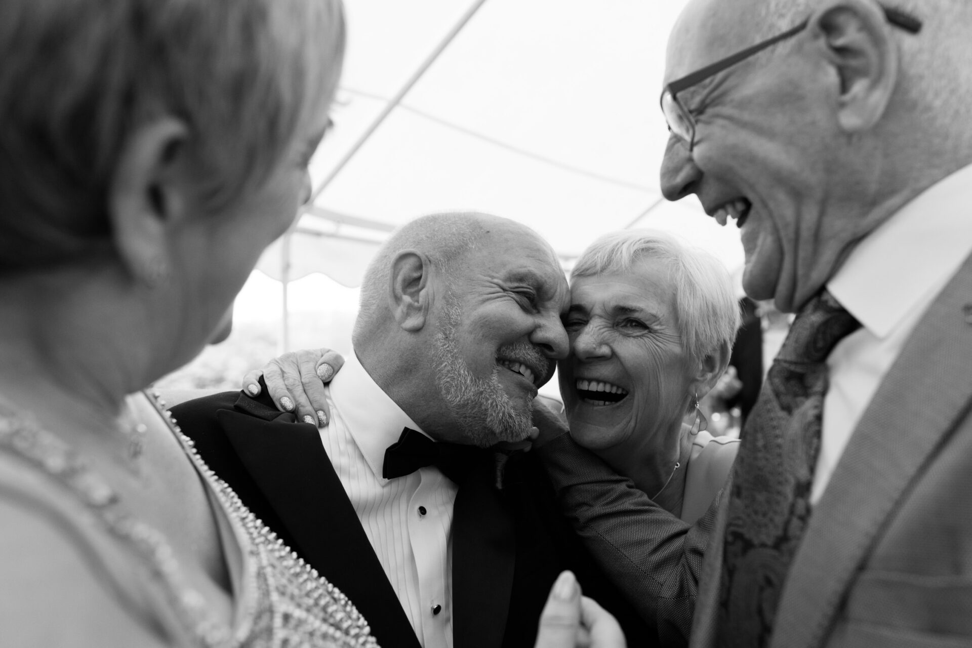 Two older couples, all dressed up in their fancy clothes, are sharing laughs and hugging each other at a nice event.