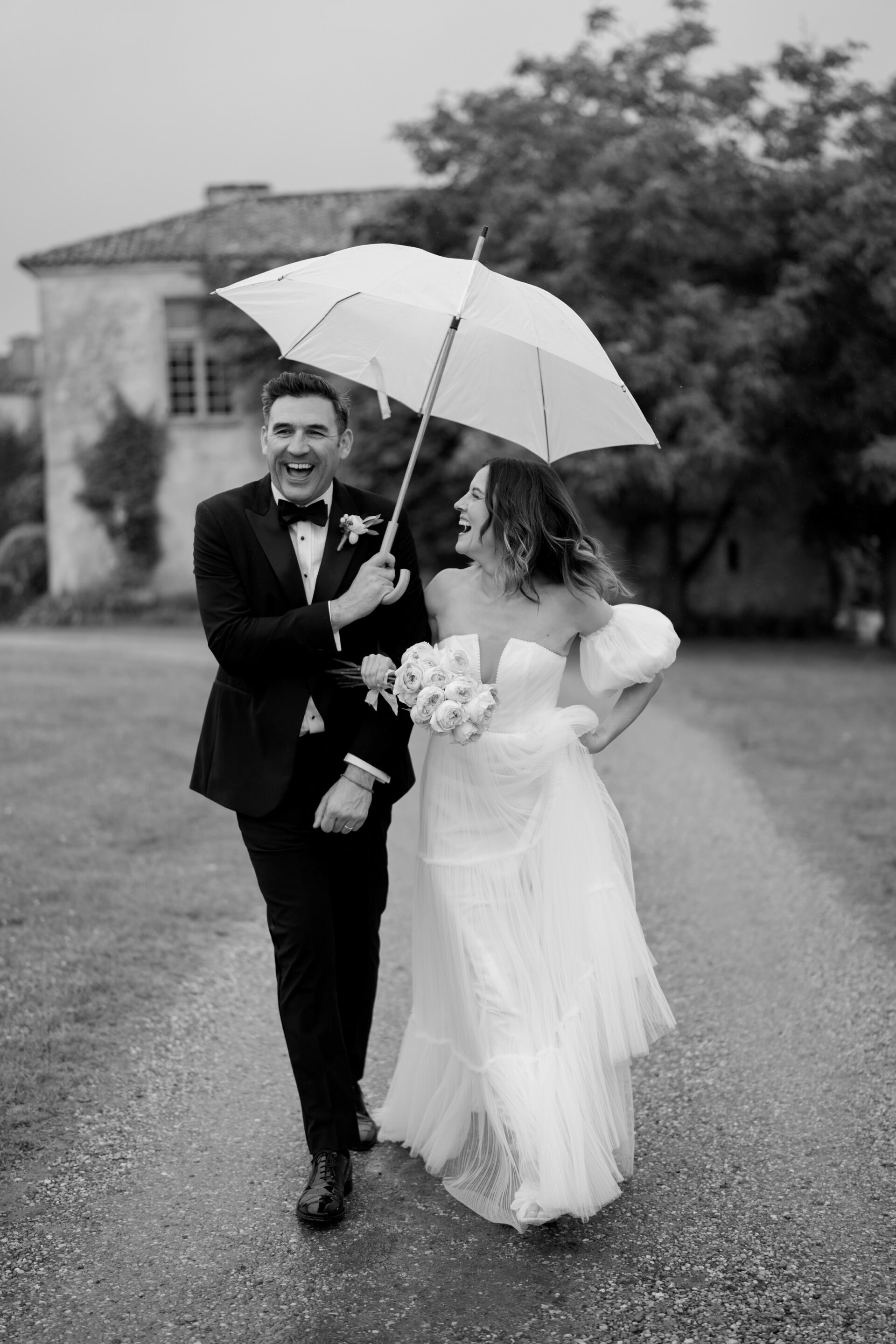 A bride and groom are smiling as they walk down a path together, sharing an umbrella. The bride is holding a bunch of flowers, and they're both wearing fancy wedding clothes.