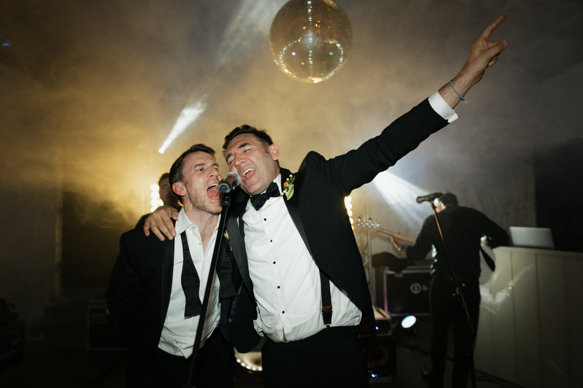 Two men in suits are enthusiastically singing into a microphone at an event, with a disco ball overhead, stage lights shining, and a musician playing in the background.