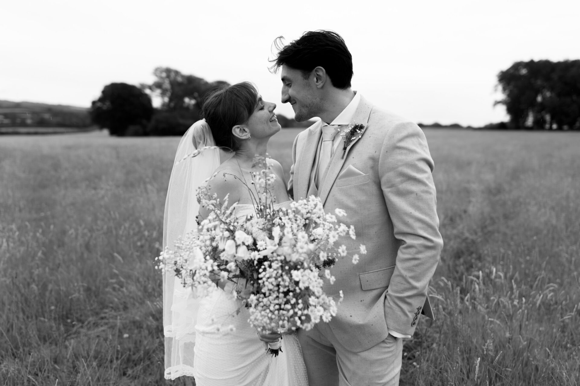 A couple dressed in wedding clothes looks at each other while standing in a field, and the bride is holding a bunch of wildflowers.