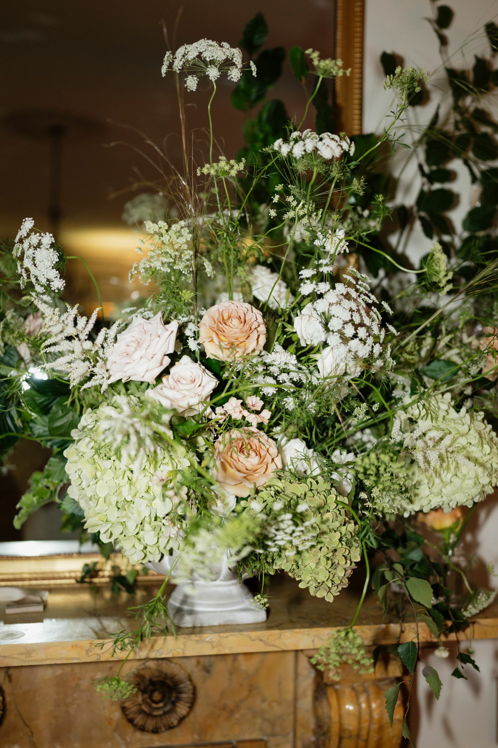 A vase with white and light pink roses, green hydrangeas, and some other green plants is sitting on a shelf above the fireplace.