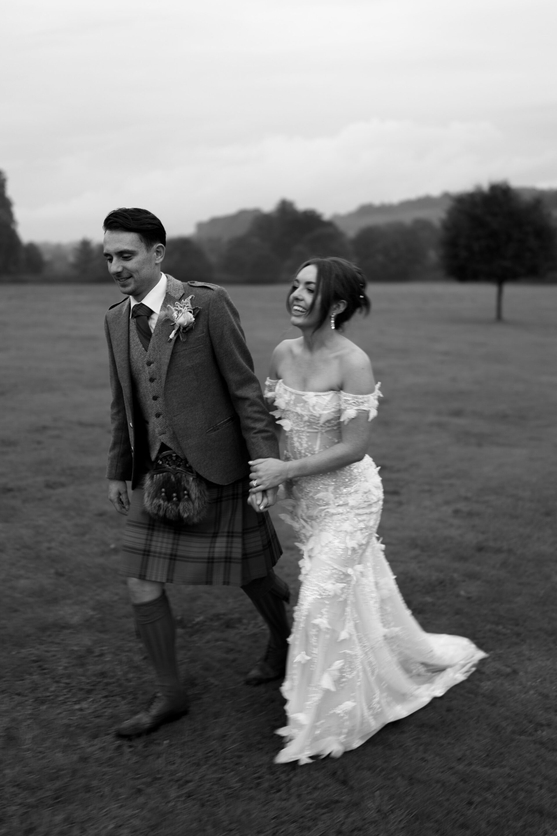 A bride wearing a lace dress and a groom dressed in a kilt are walking hand in hand across a grassy field.