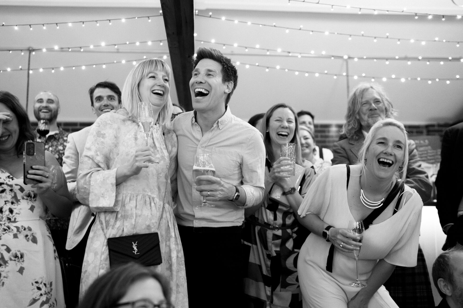 A bunch of people are laughing and having drinks at a celebration, with string lights hanging above them.