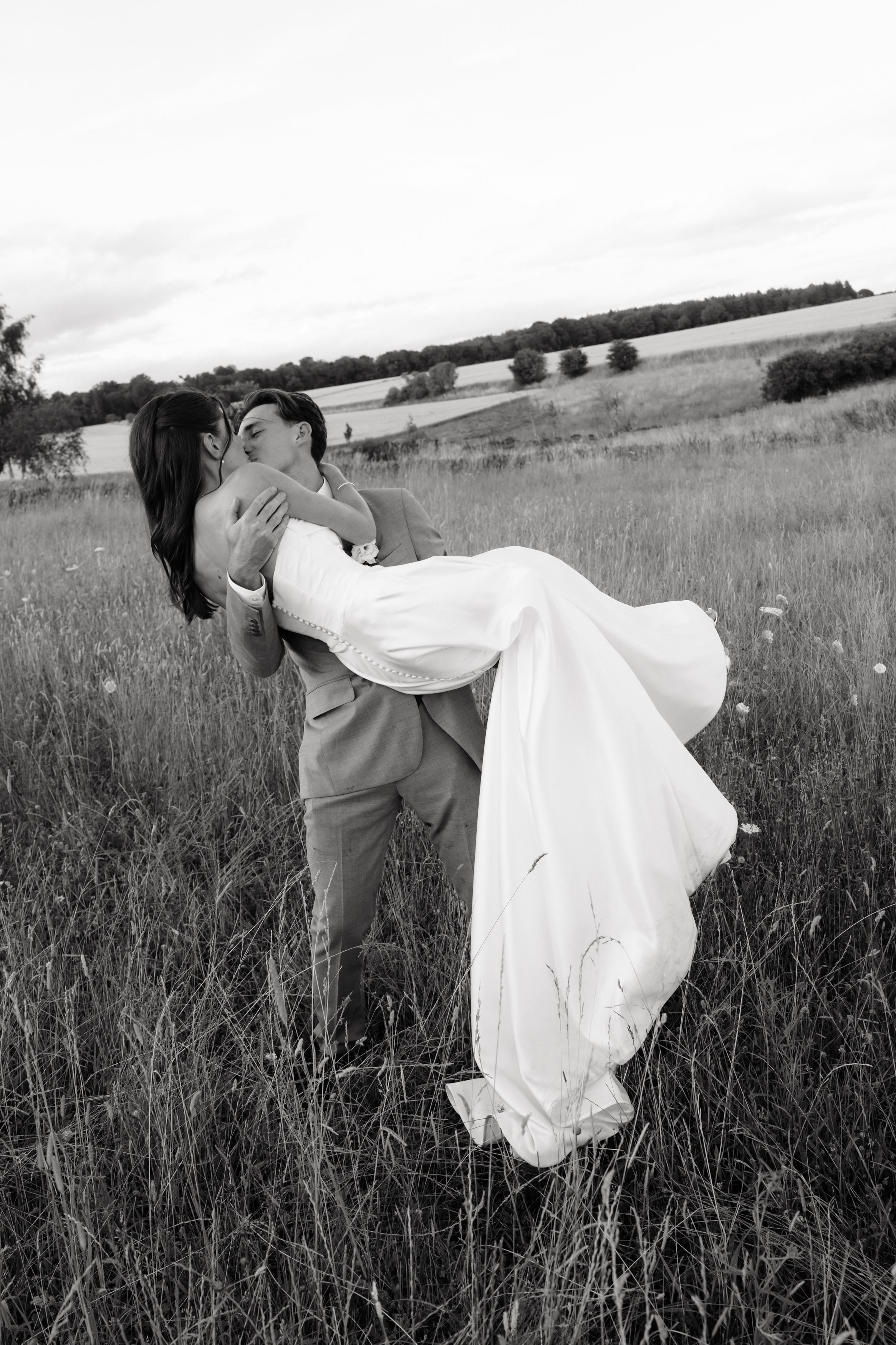 A groom is carrying his bride, and they’re kissing as they walk through a grassy field. They’re outside, with trees and water behind them.