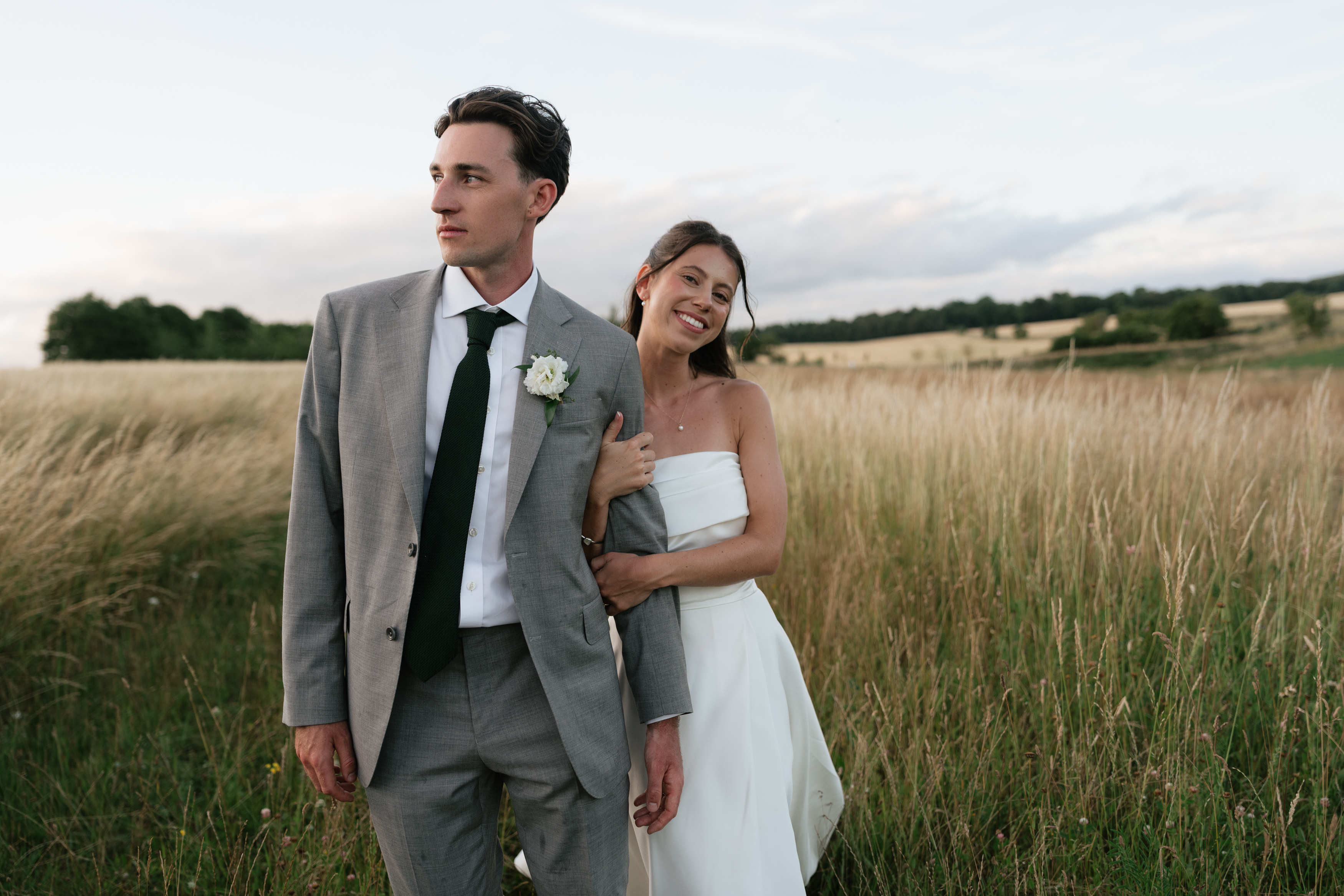 A woman in a white dress is smiling with her arm linked to a man in a grey suit as they pose together in a grassy field.