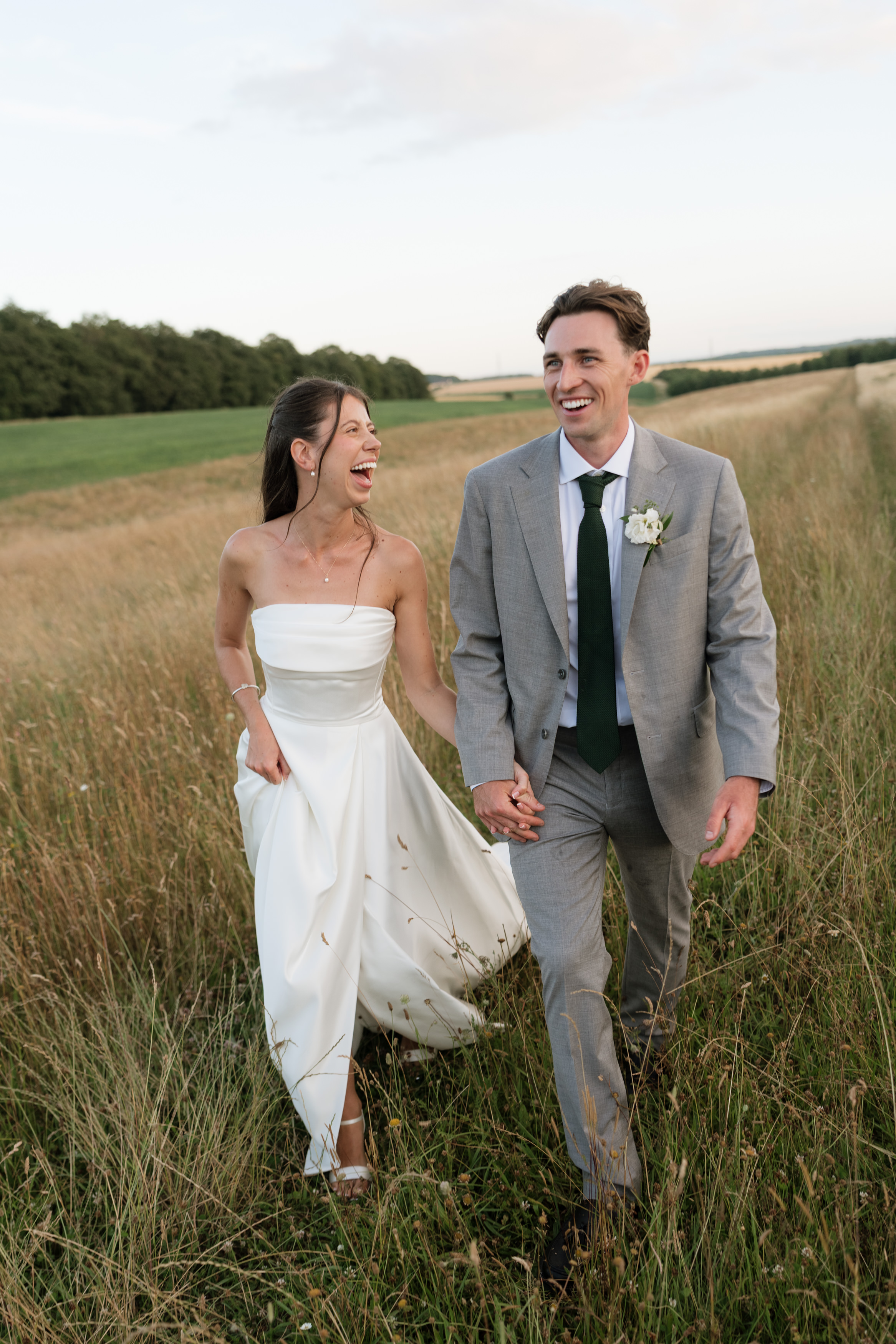 A woman wearing a white wedding dress and a man in a grey suit are walking together, holding hands in a grassy field. They’re both smiling and looking at each other.