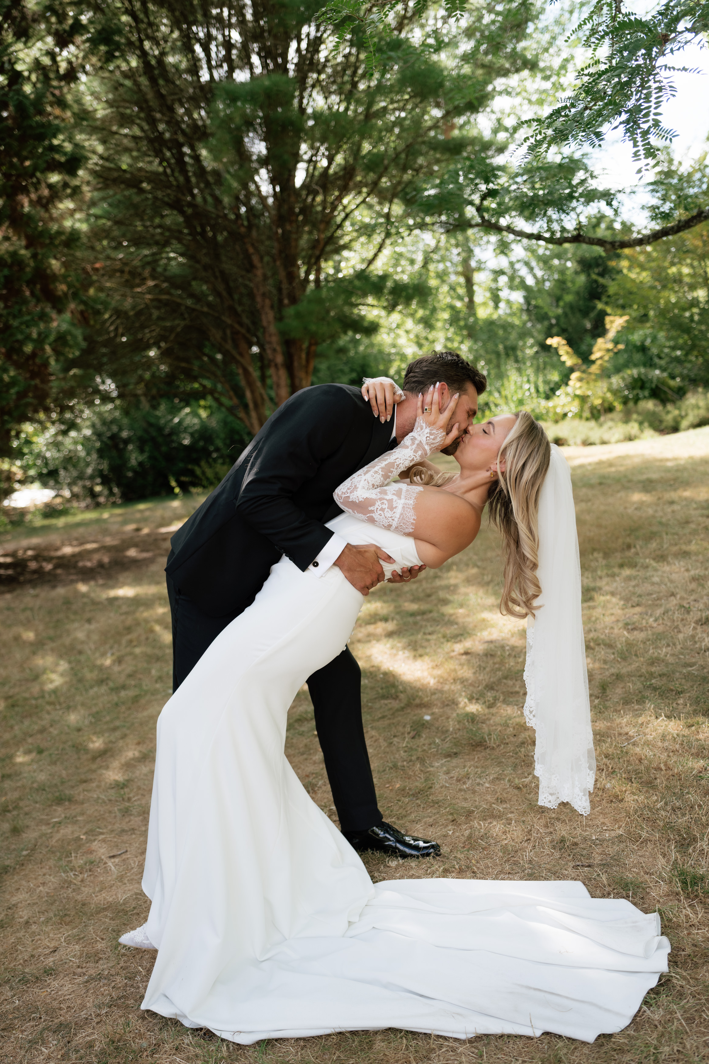 A groom wearing a black suit dips and kisses his bride, who’s in a white dress and veil, outside on the grass with trees around them.