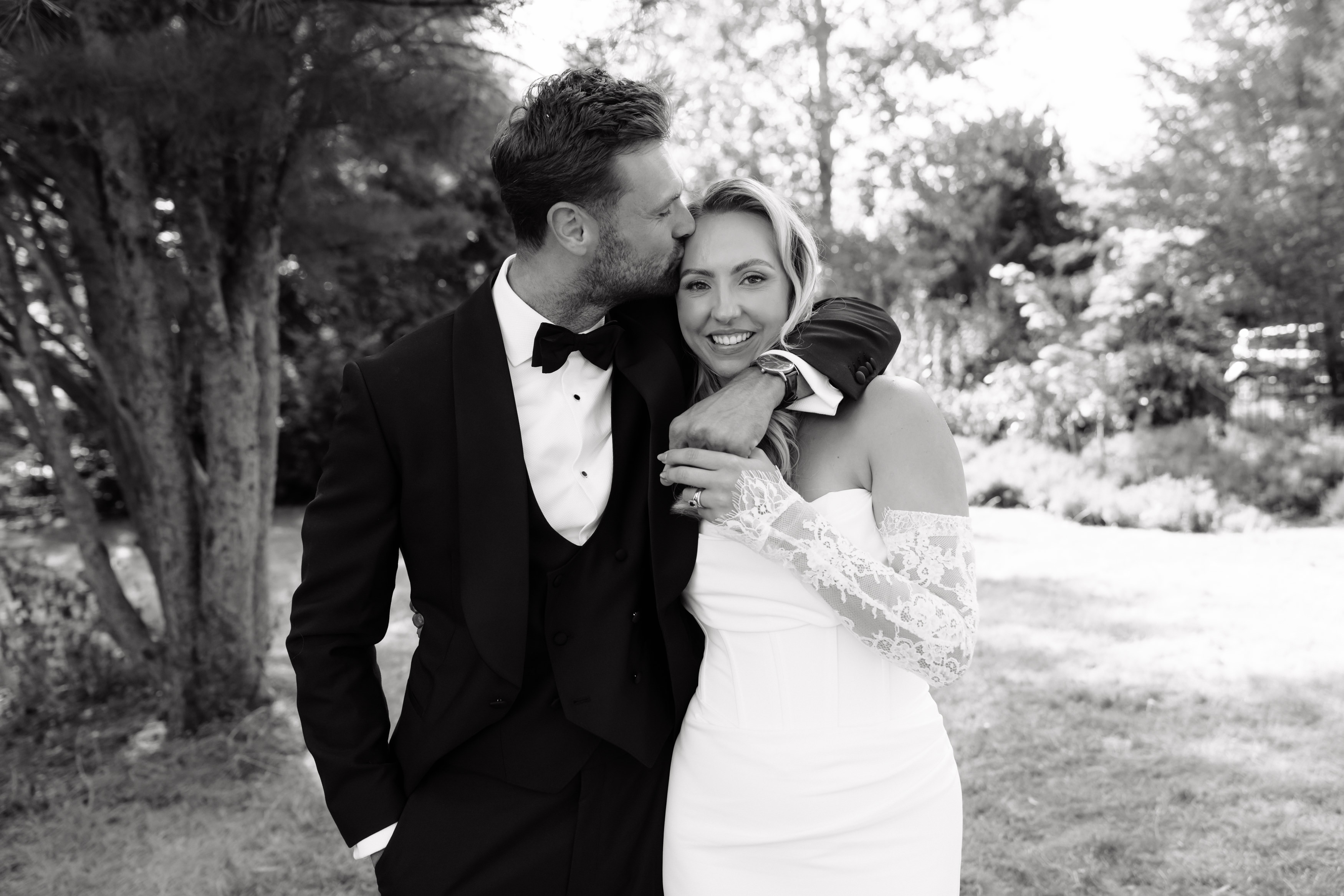 A man wearing a dinner jacket gives a head kiss to a smiling woman in a white wedding dress while they hug and pose together outside in a garden.