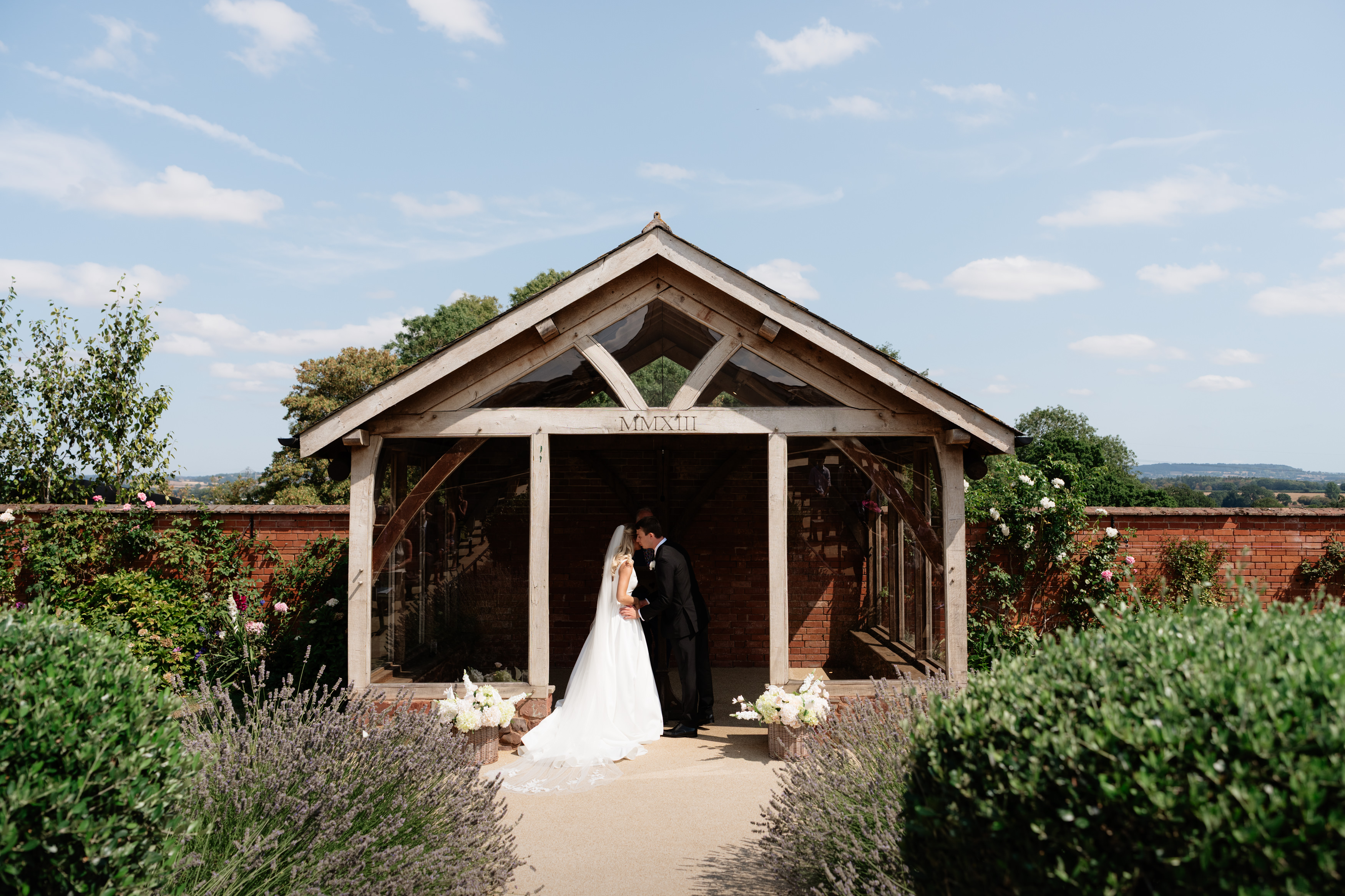 A bride and groom share a kiss under a wooden pavilion outside, with lots of plants and potted flowers around them on a sunny day.