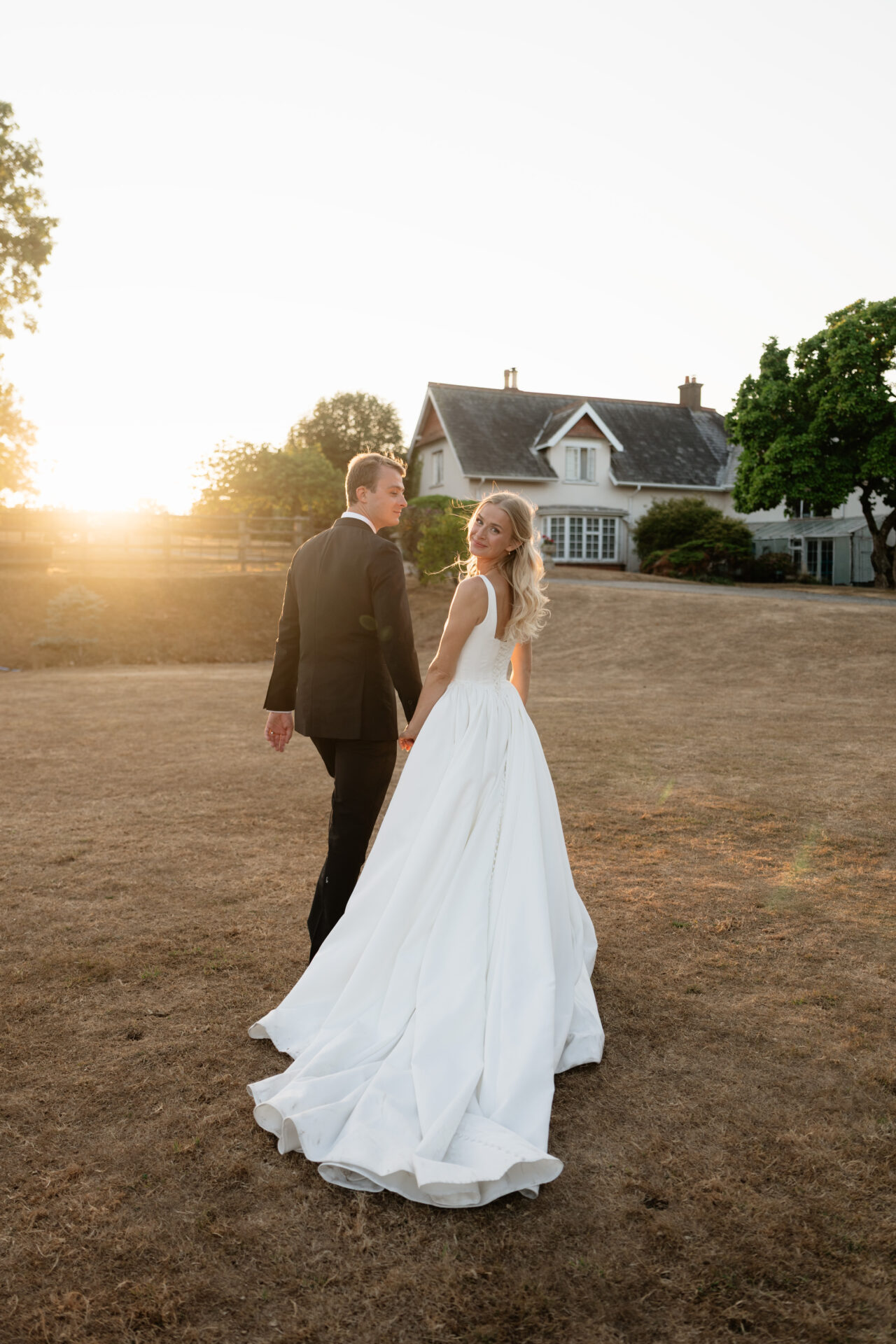 A bride in a white dress and a groom in a black suit are holding hands as they walk outside a house during sunset.