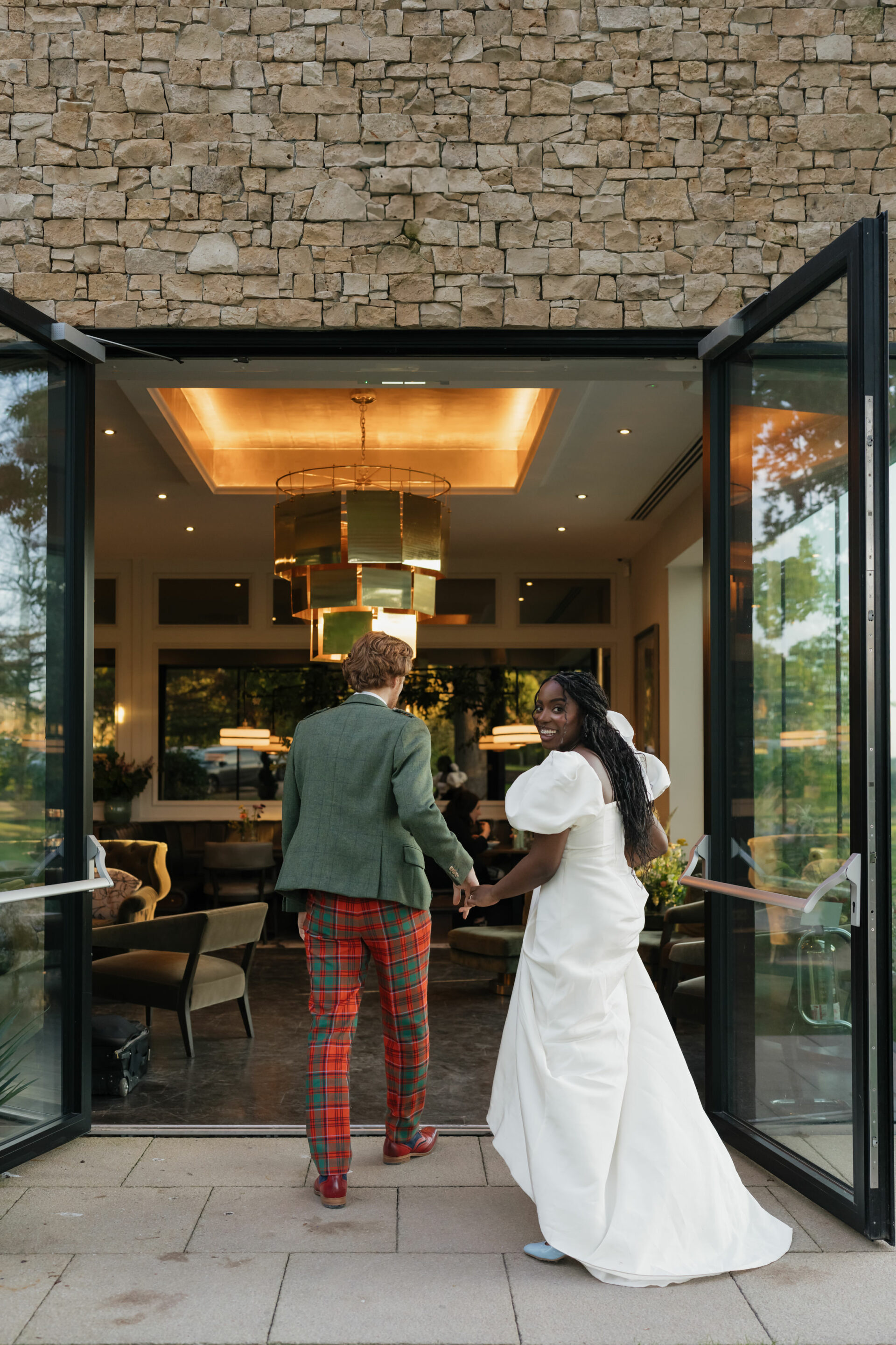 A couple walks into a building with big glass doors, one wearing a white wedding dress and the other in a green jacket with plaid pants.