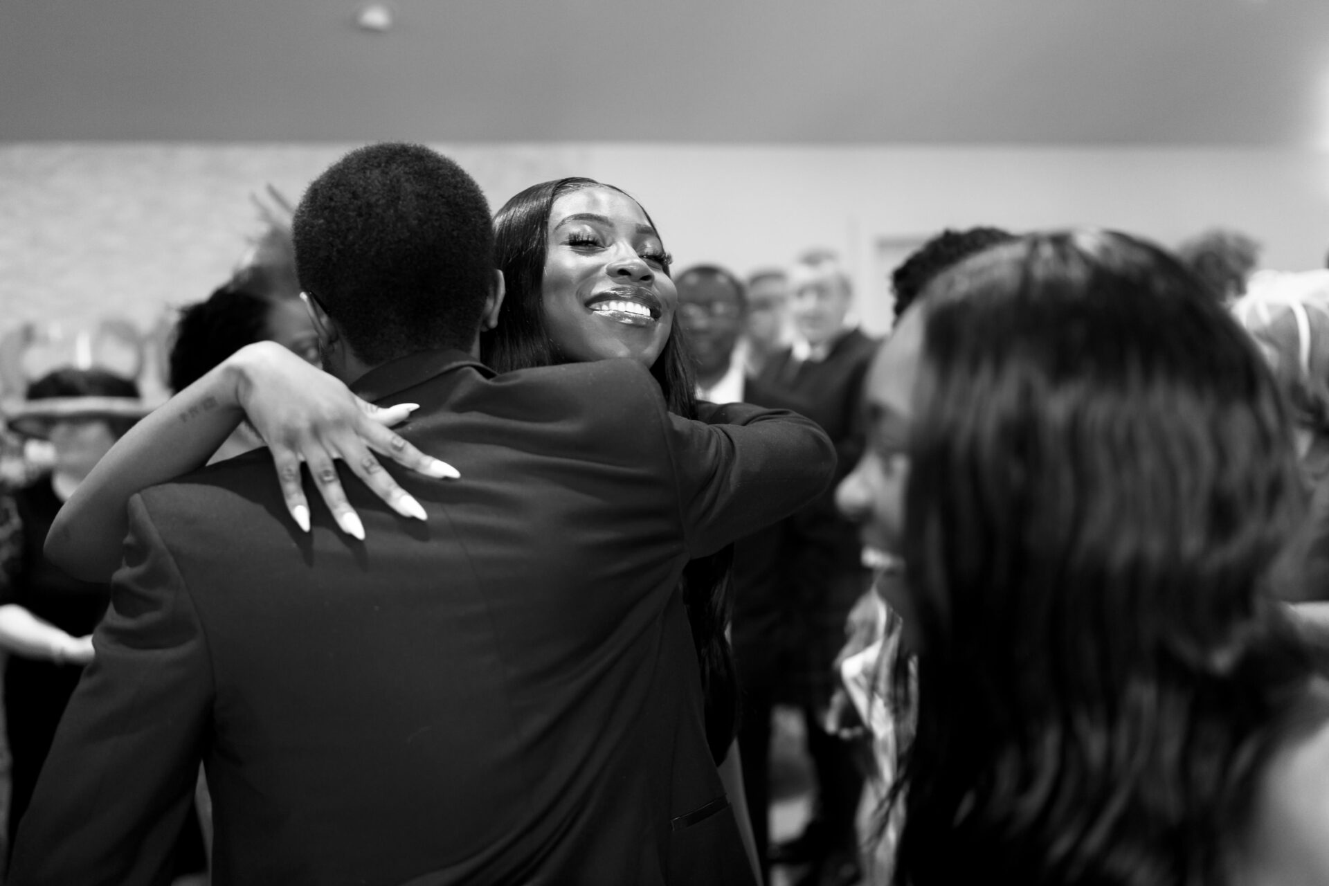 A woman happily hugs a man in a busy room, celebrating something together. In the background, other people are smiling and chatting with each other.