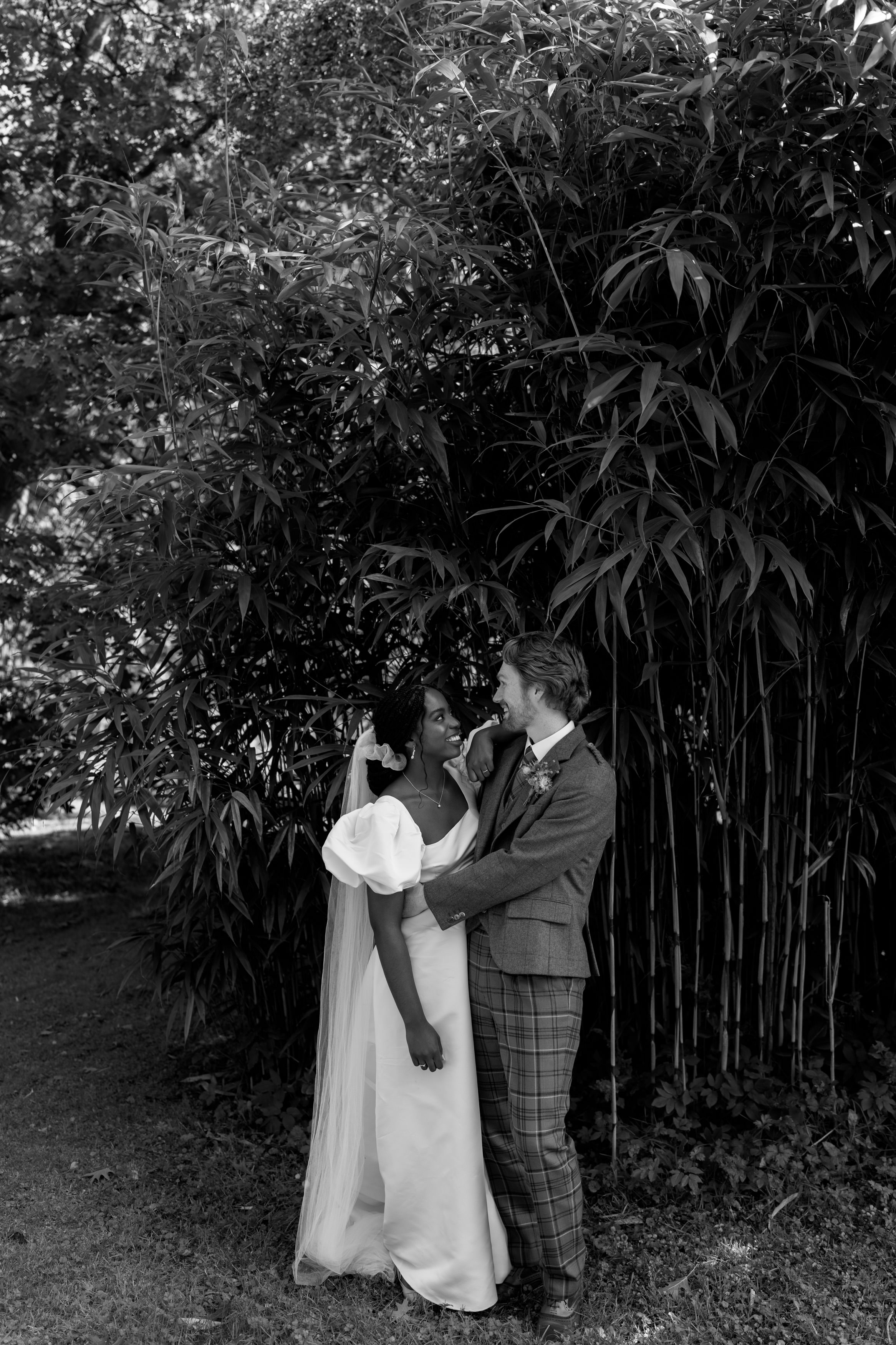 A couple is standing close together in front of tall bamboo plants; the woman is wearing a dress, and the man is in a suit. They're sharing a sweet moment.