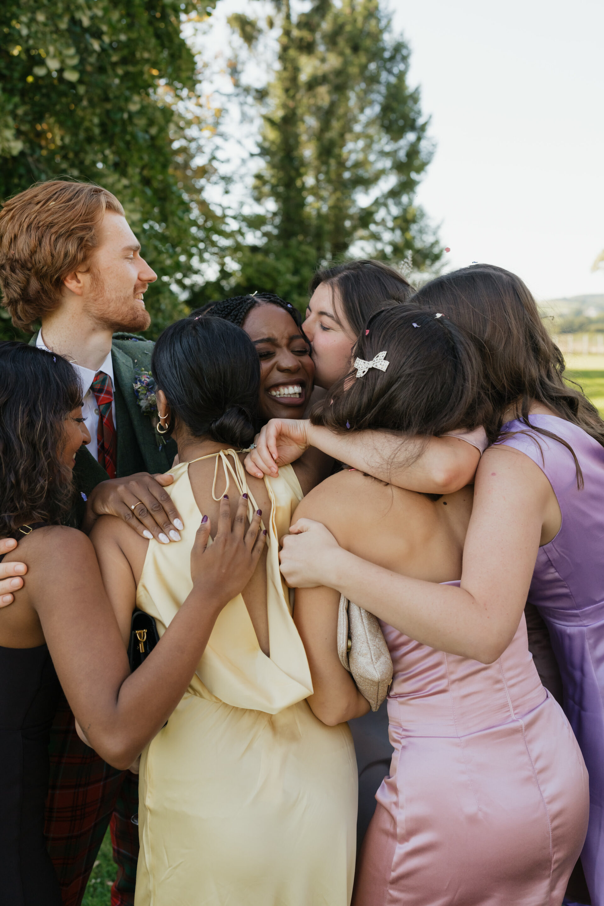 Seven people in fancy clothes are happily hugging and smiling outside, with trees behind them.