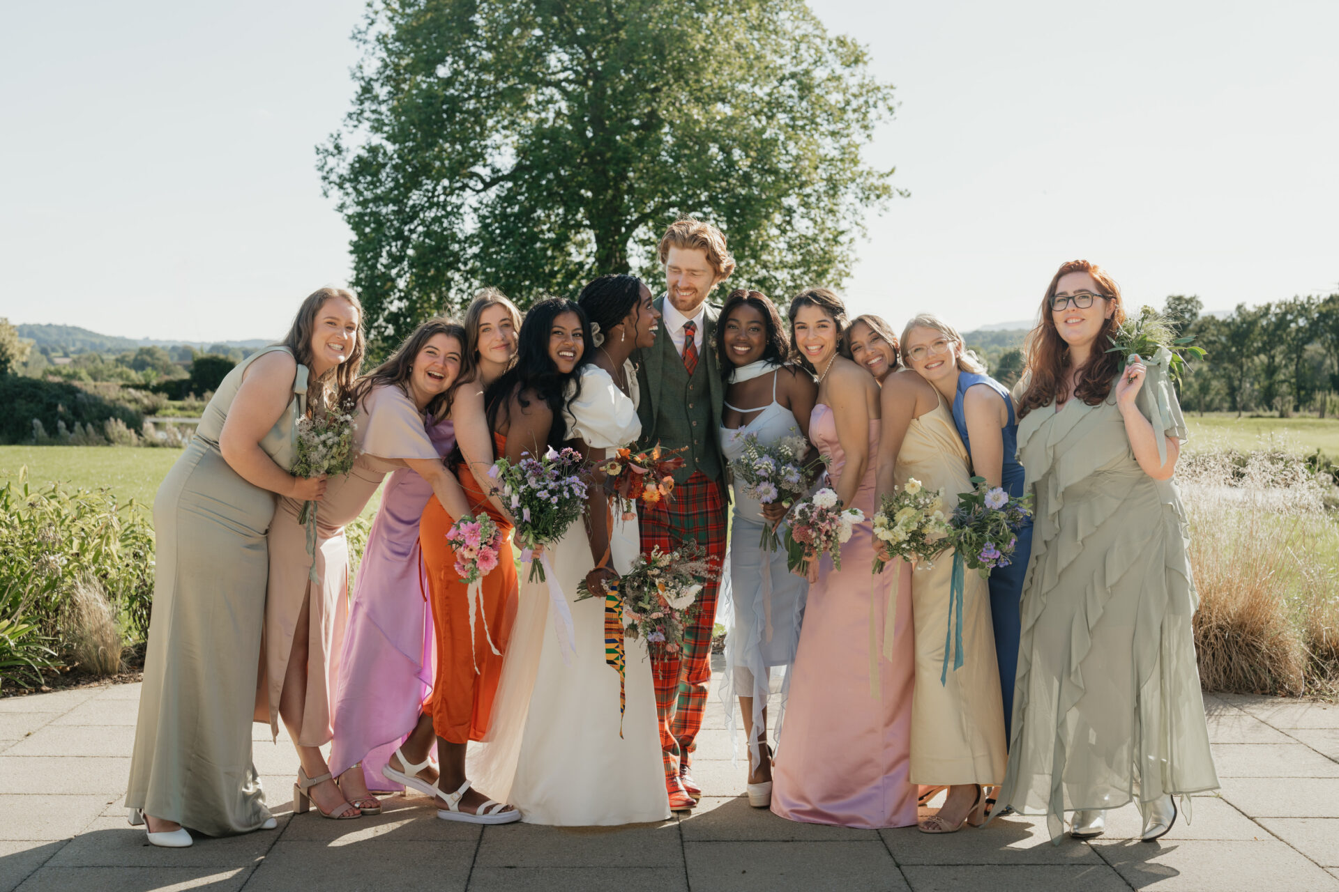 A group of people are standing together outside, smiling and holding flowers. One person is in a wedding dress and another is wearing a suit.
