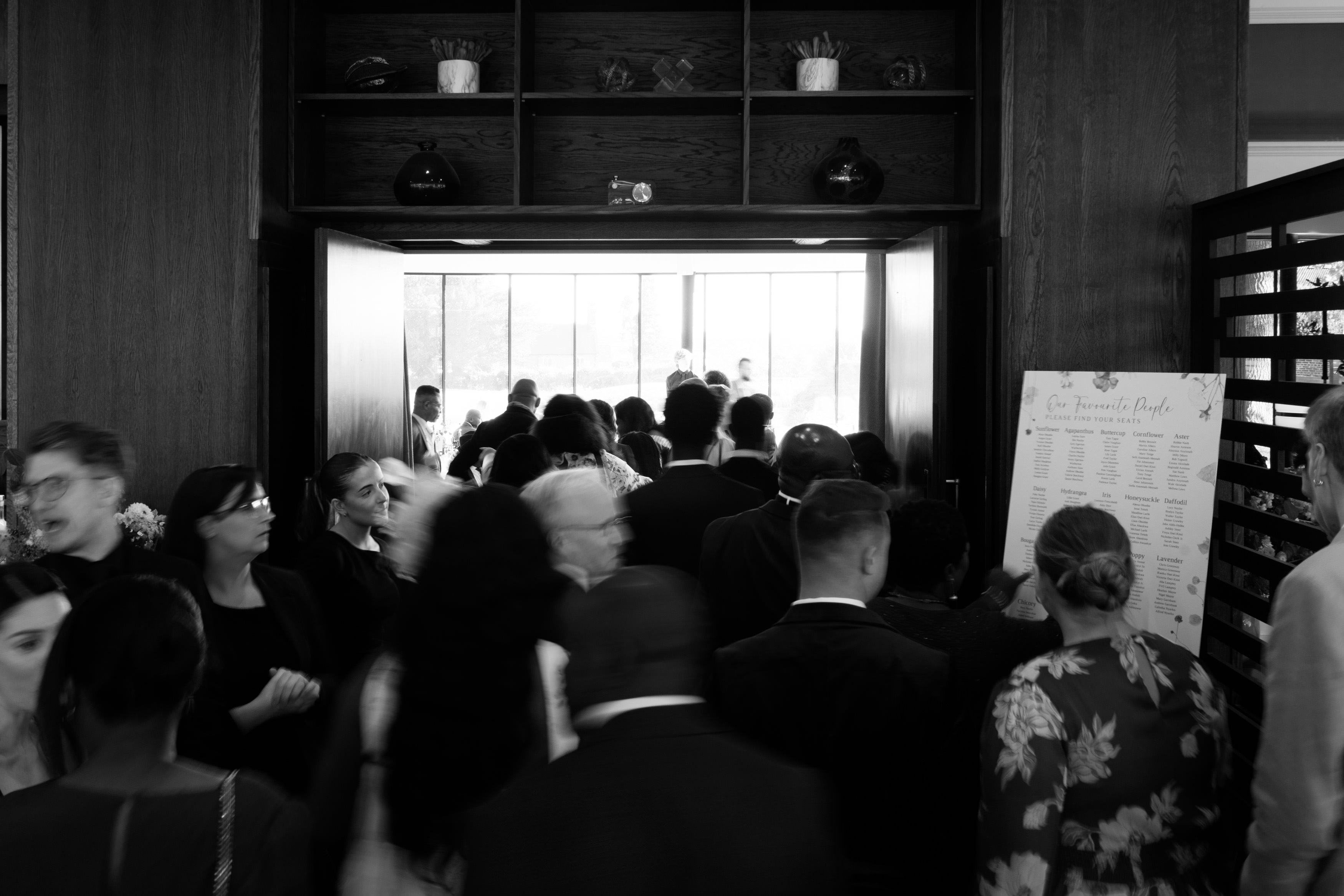 A group of people dressed up nicely walks into a place through big double doors. There's a seating chart on the right side.