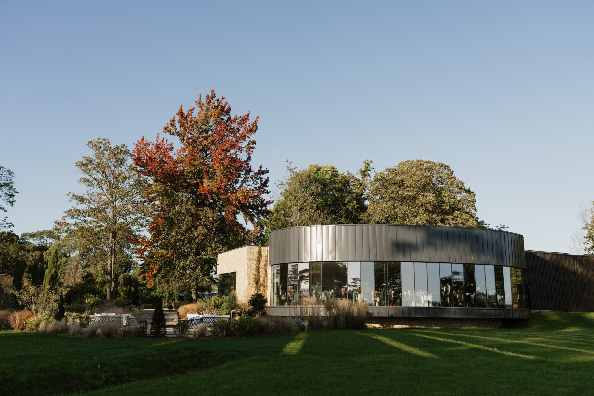 A round building with lots of big windows sits in the middle of a grassy area with trees. One tree has bright autumn-colored leaves, and the sky is clear and blue.