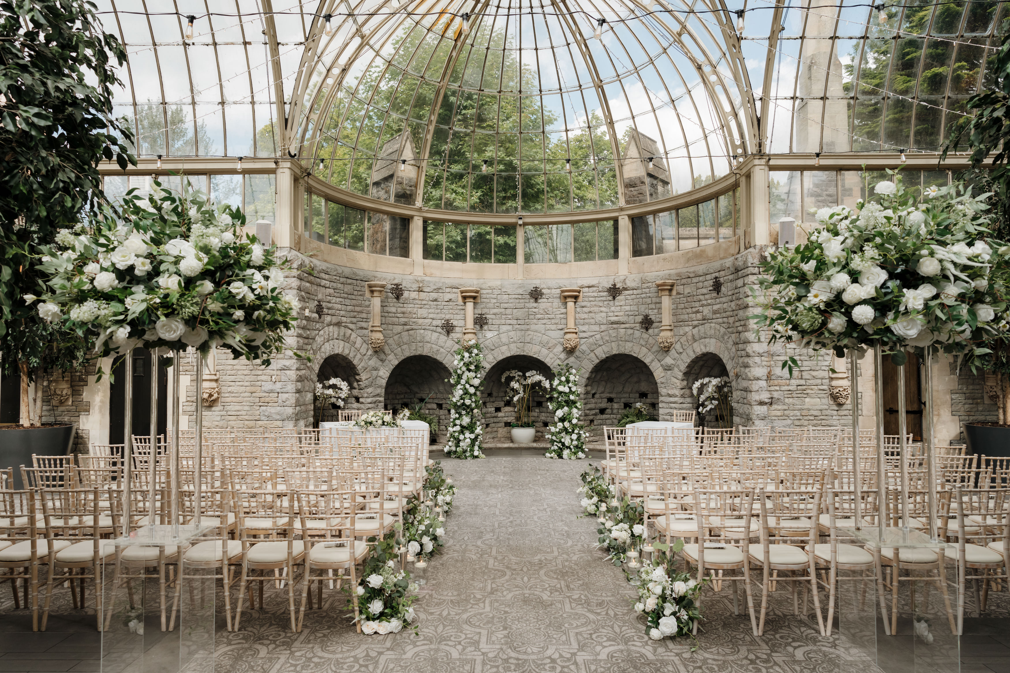 A stone building with a glass roof filled with rows of chairs, white flowers, and an archway all set up for a wedding.