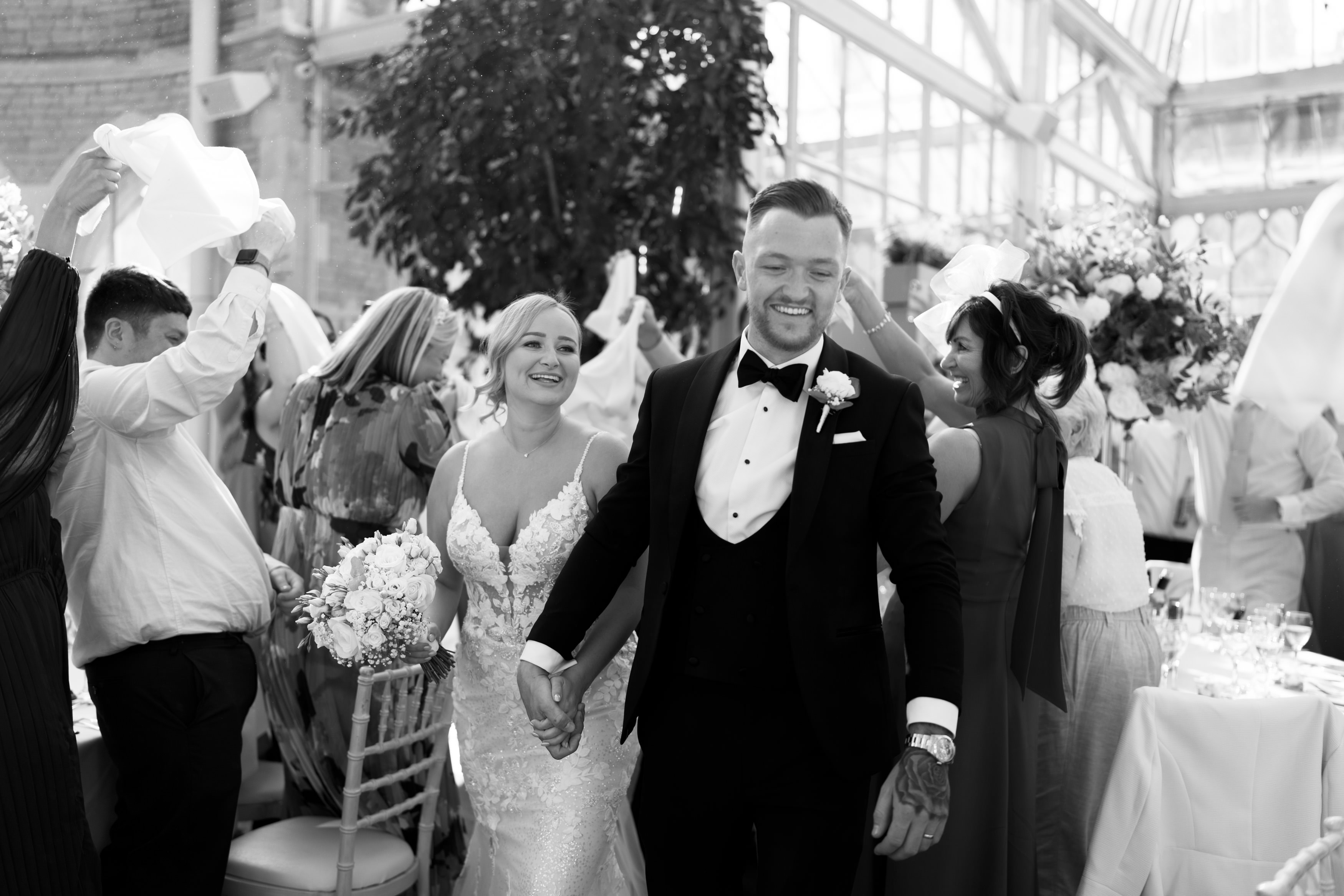 A bride and groom walk hand in hand, smiling, as they move through a happy crowd that's waving napkins in an indoor space with big windows.