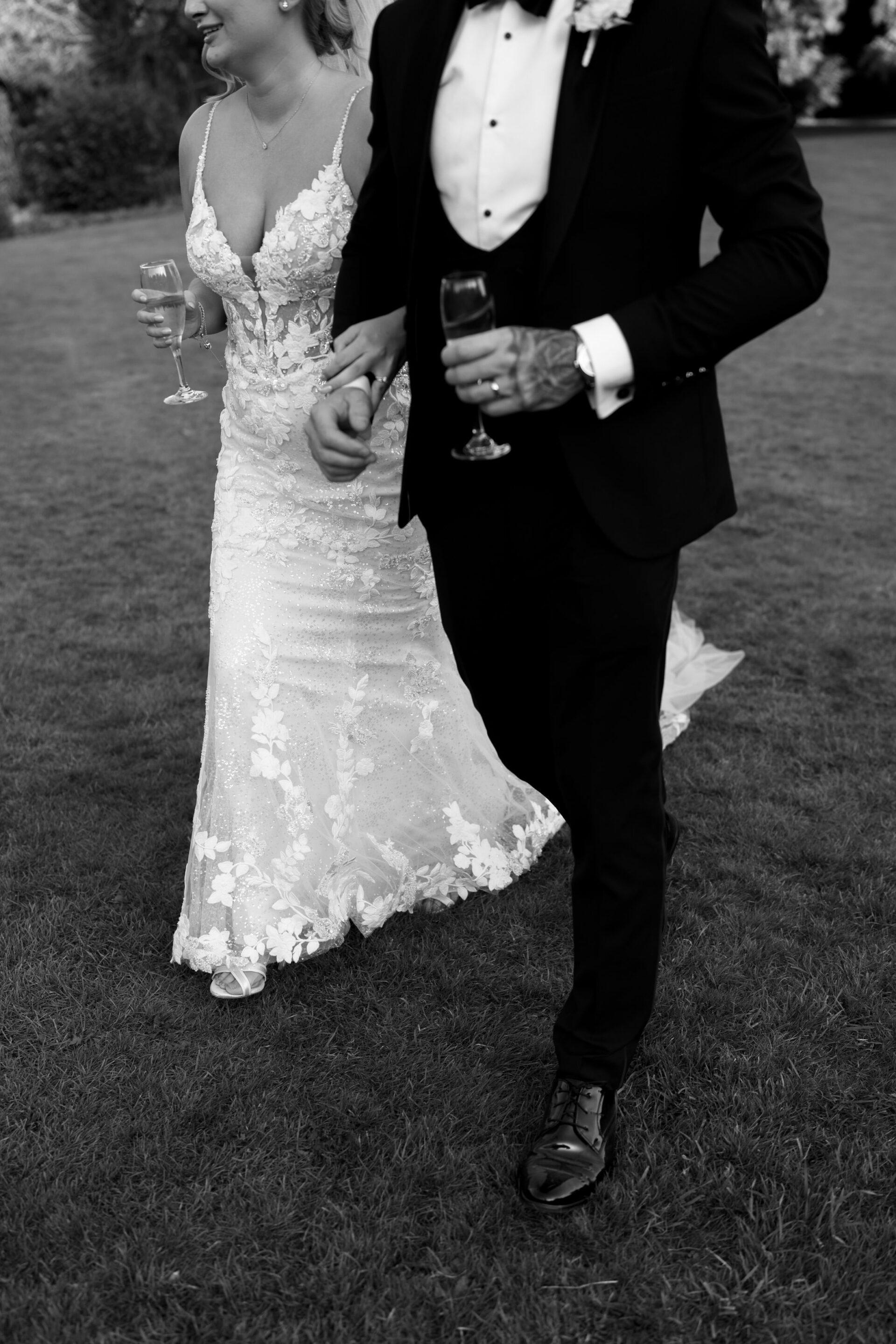 A dressed-up couple walks hand in hand on the grass while holding champagne glasses.