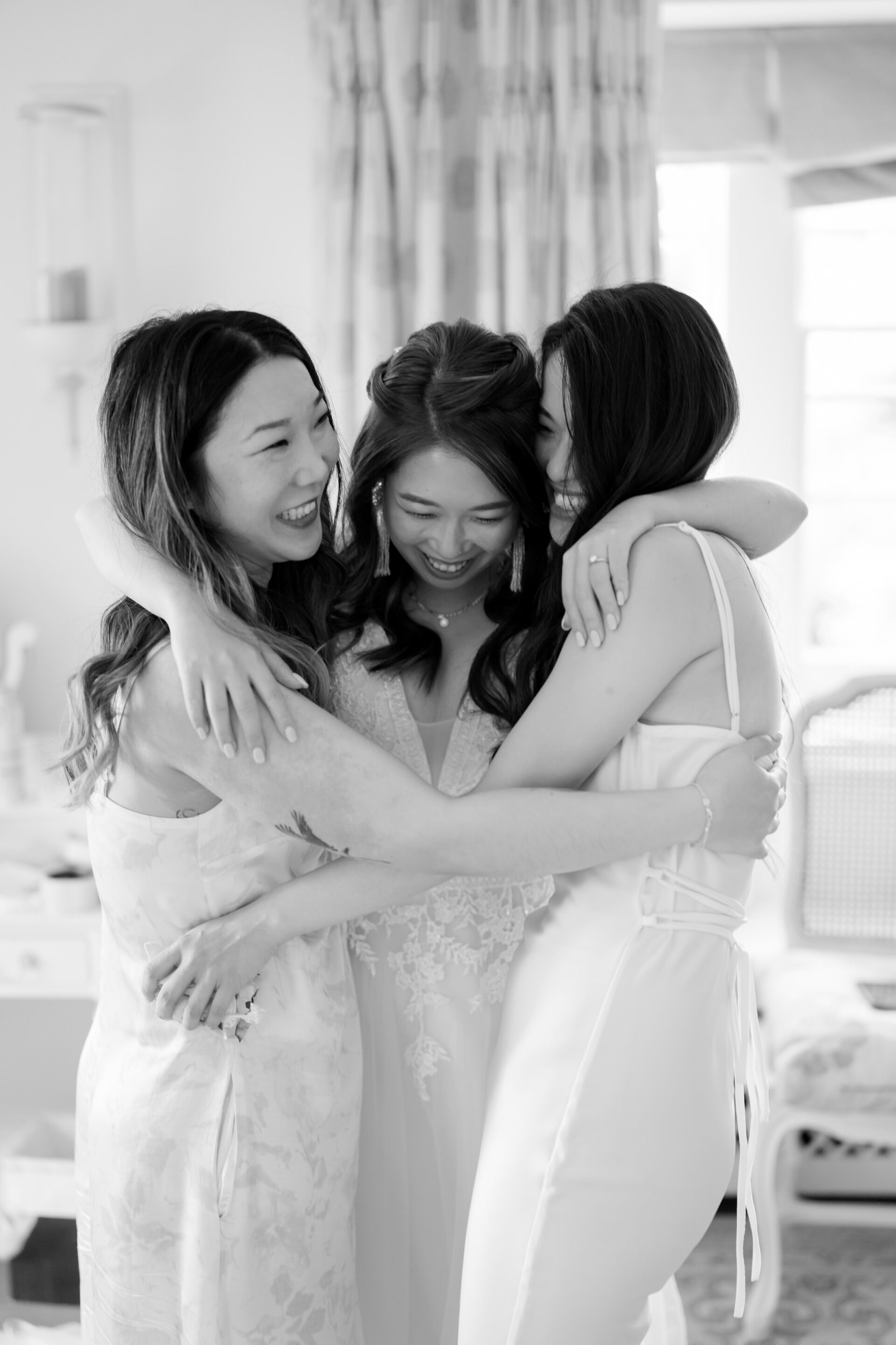 Three women in dresses happily hug each other in a bright room.