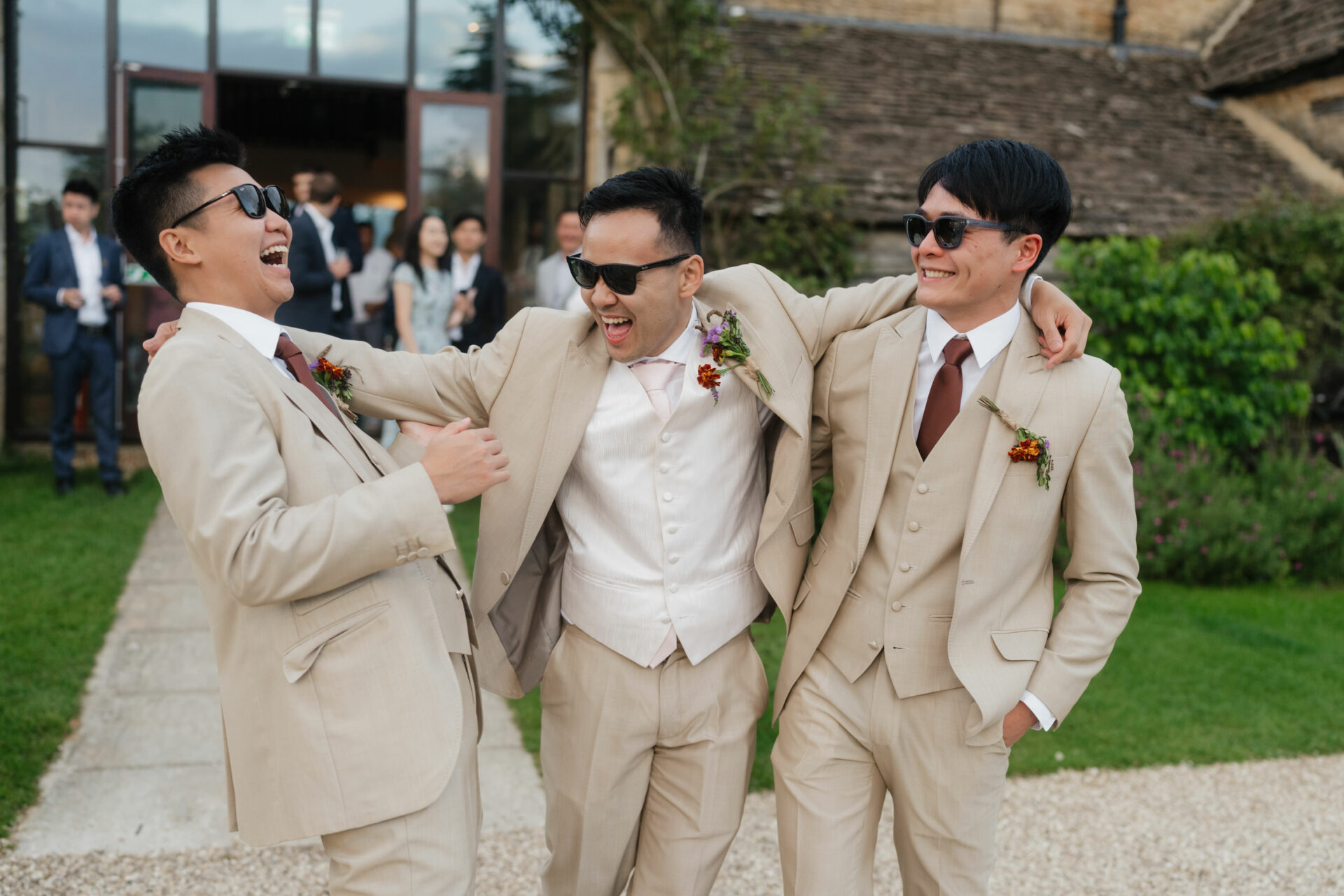 Three guys in beige suits and sunglasses are laughing together outside on a sunny day, with a bunch of people in the background.