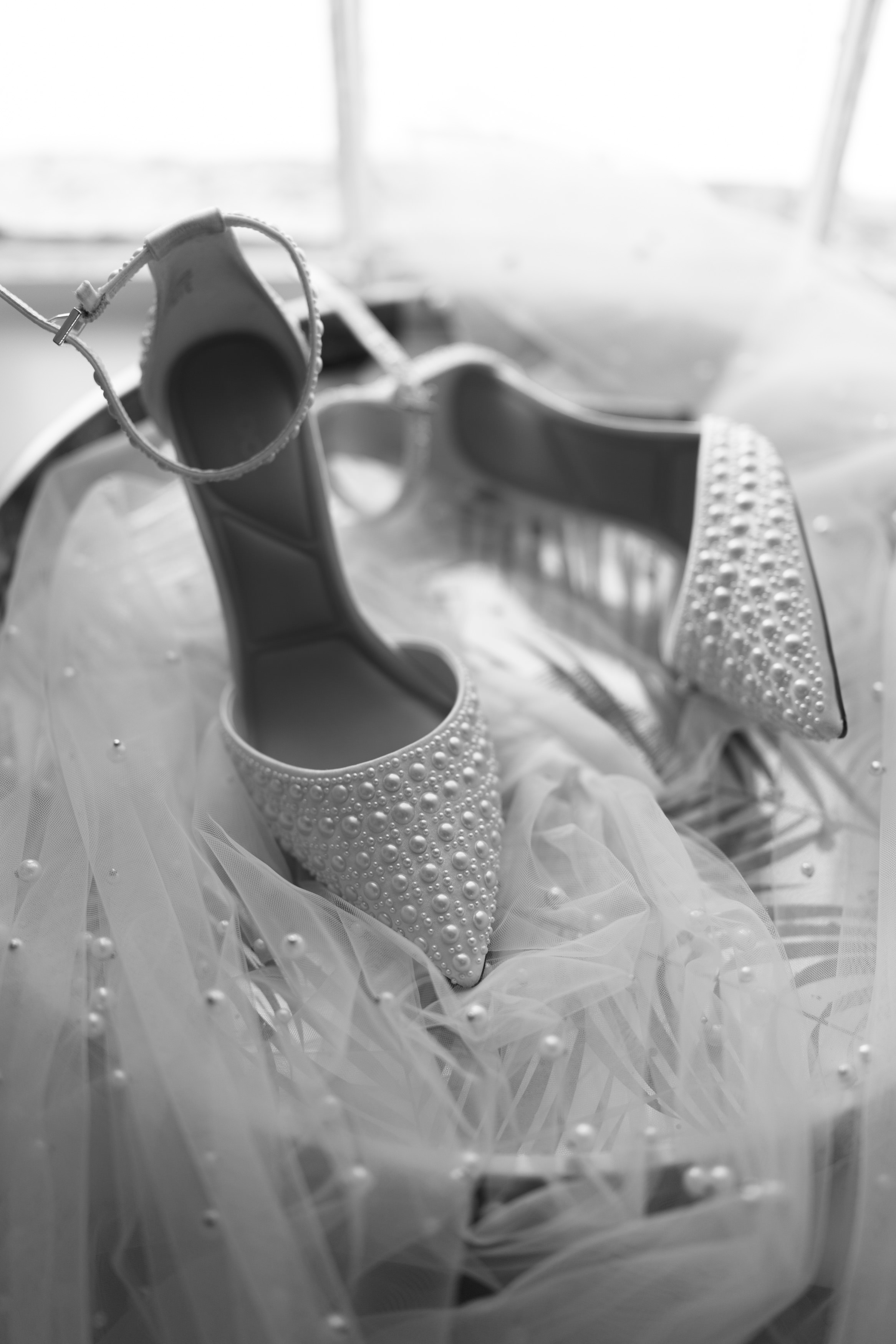 A black and white photo of stylish pointy high heels decorated with pearls, sitting on a light, see-through fabric.