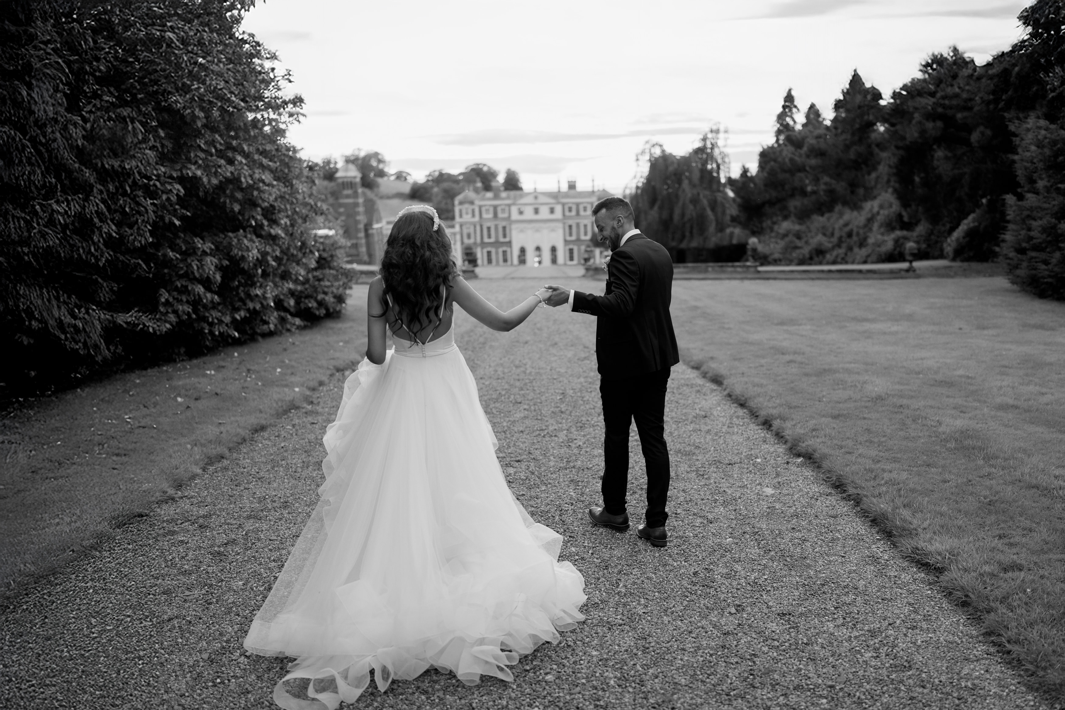 A bride and groom are walking together hand in hand on a gravel path. There's a big building behind them, and trees are lining both sides of the path.