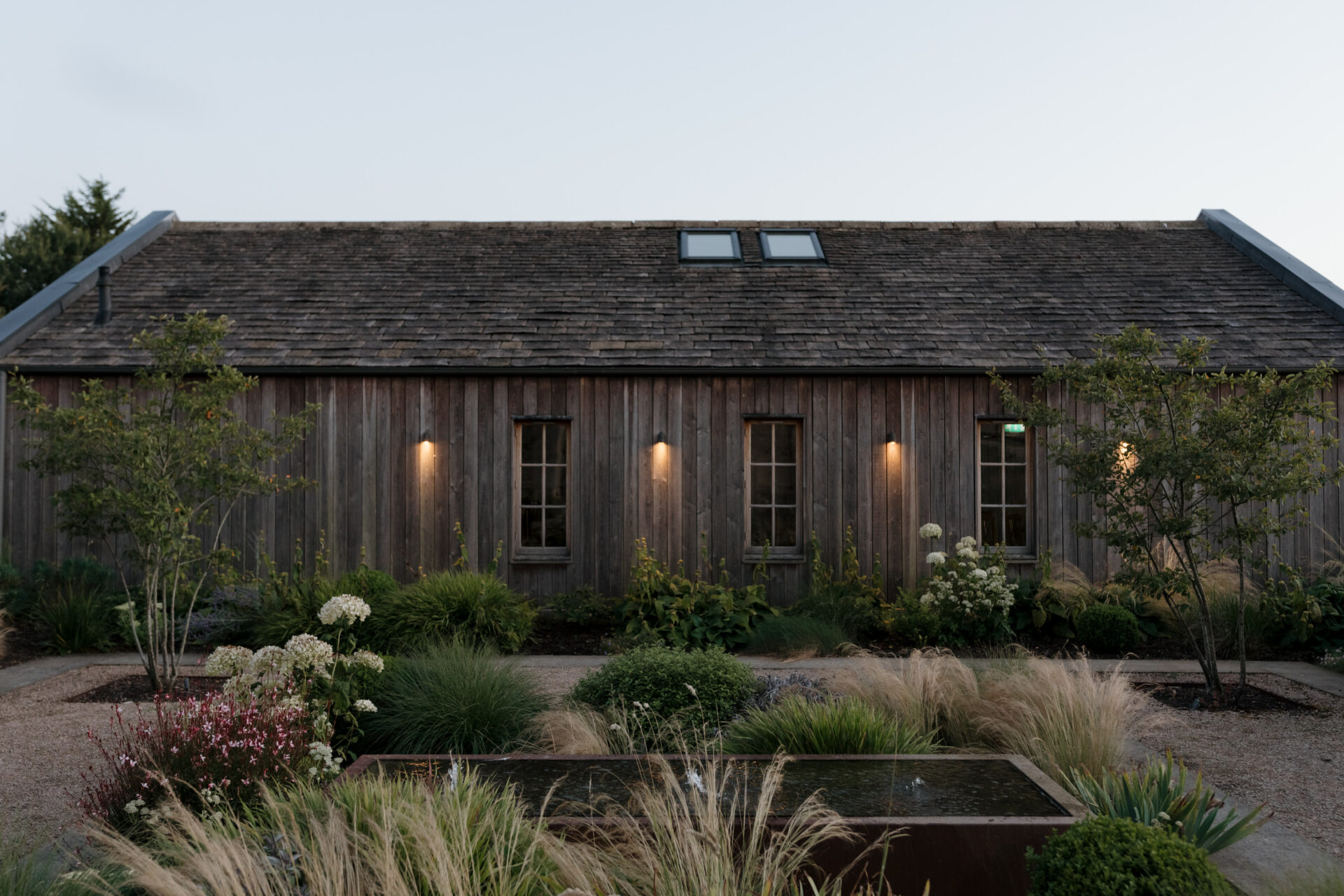 A wooden house with three windows is surrounded by a landscaped garden full of grasses and flowers. The house has four outdoor lights, and the roof features two skylights.