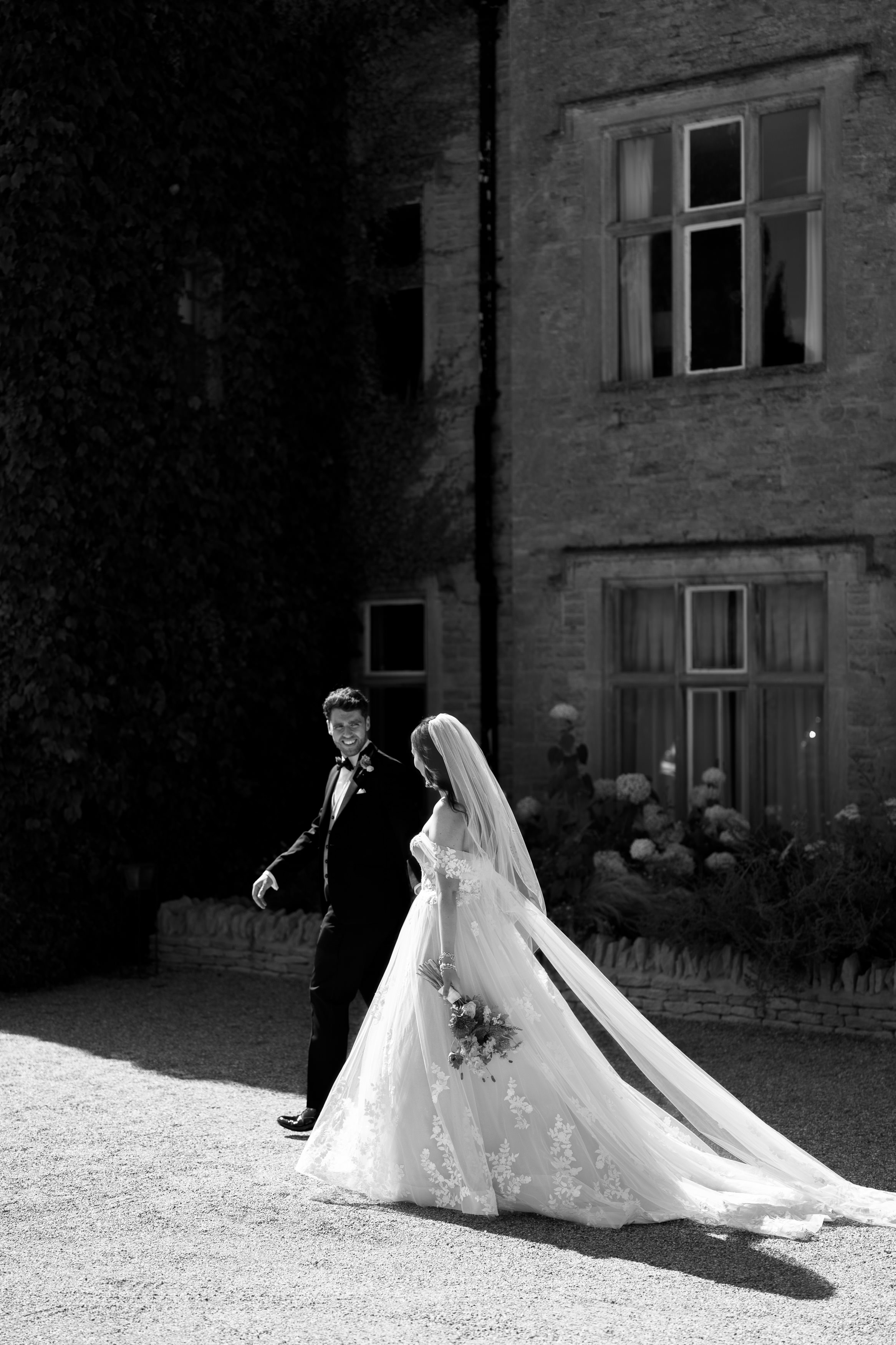 A bride in a long, flowing dress holding a bouquet walks with the groom outside a stone building.