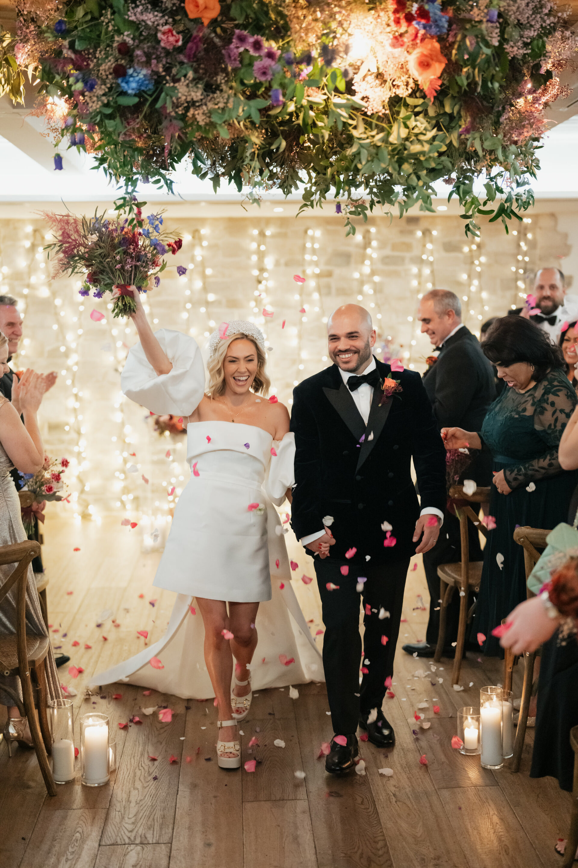 A happy couple walks down the aisle at their wedding while guests clap and flower petals fall around them. They pass under an arch decorated with flowers and lights are shining in the background.