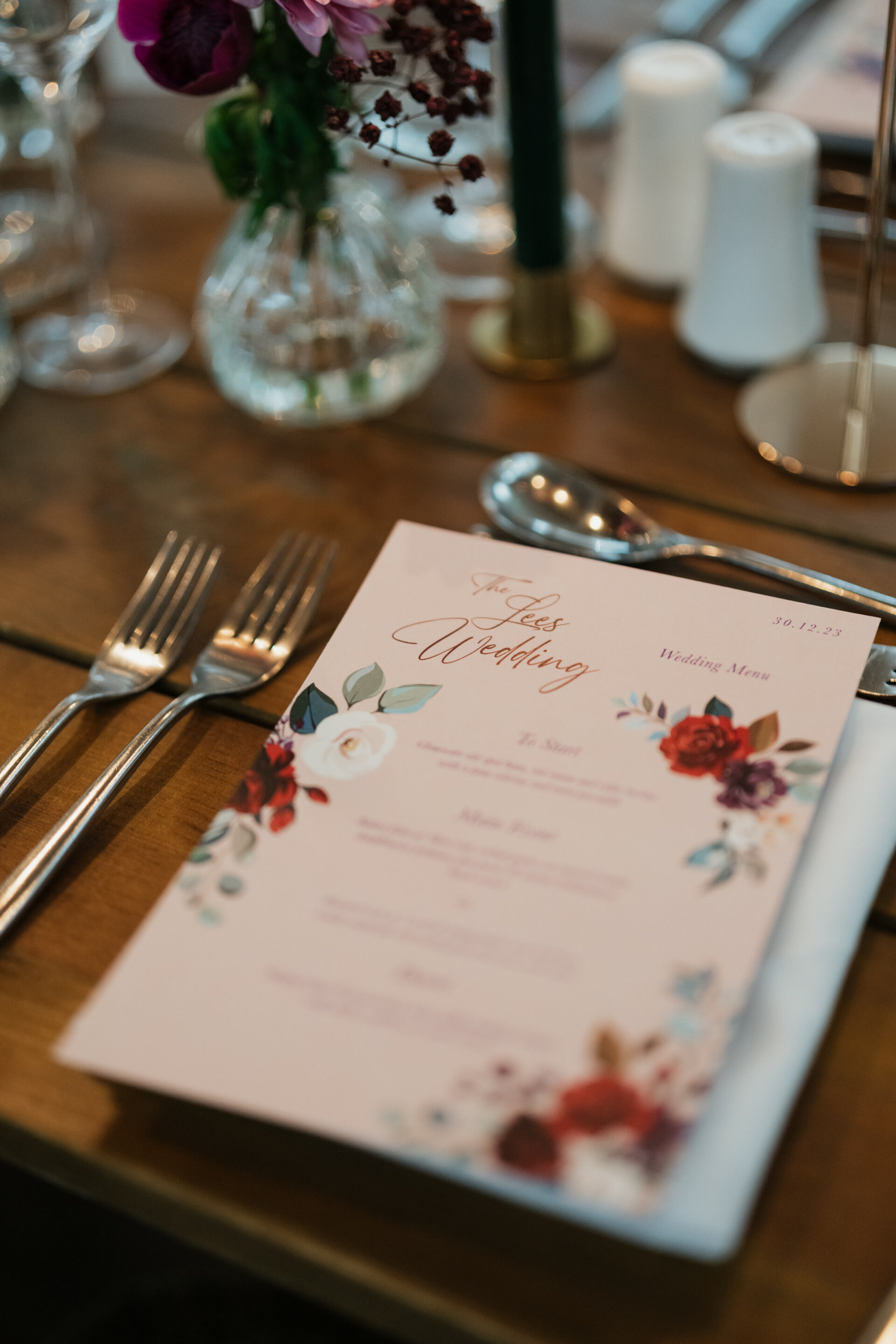 A wedding menu with a floral design is placed on the table between a fork and spoon. There are glasses and a flower arrangement in the background.